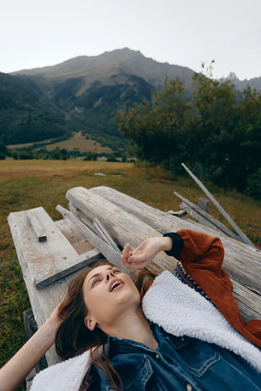 woman on wooden bench in meadow outdoors with mountains in background, nature scene showing relaxation and casual jacket, portrait of young female reclined enjoying peaceful countryside landscape