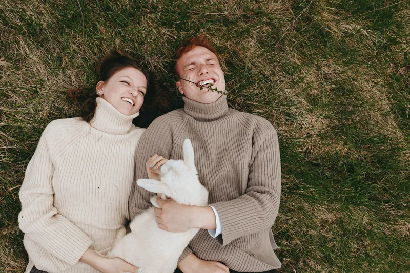 A couple lies on grass with a playful goat kid nestled between them, wearing sweaters in a sunlit meadow. A lifestyle moment that highlights affection, companionship, and rustic charm of a farm scene.