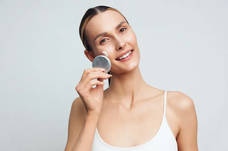 Woman shows beauty skincare serum holding a glass vial near her face studio spa portrait clean white background calm smile hygiene wellness glow hydration care