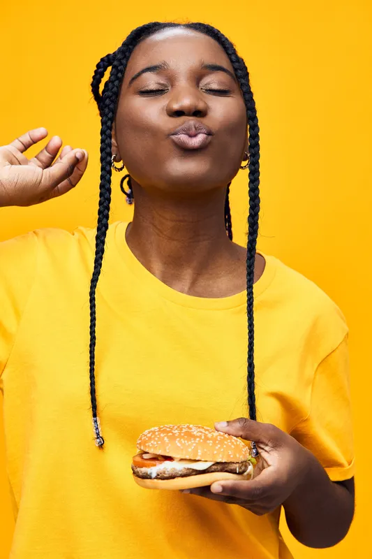 Happy young woman with braids enjoying a delicious burger against a bright yellow background, showcasing joy and satisfaction in healthy eating