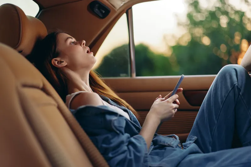 Young woman relaxing in a car, holding a phone, dressed casually, with a peaceful expression, surrounded by warm evening light and greenery.