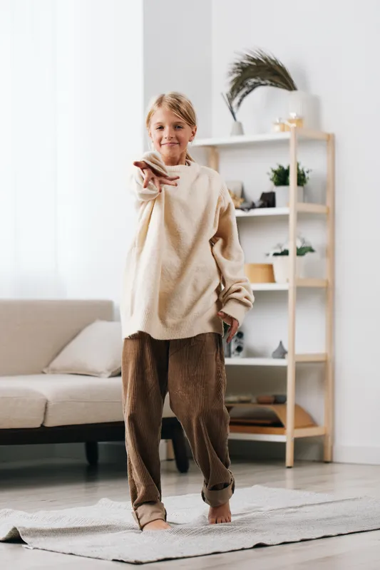 A warm, stylish woman stands in a bright living room wearing an oversized beige sweater and relaxed brown pants, pointing playfully toward the camera.