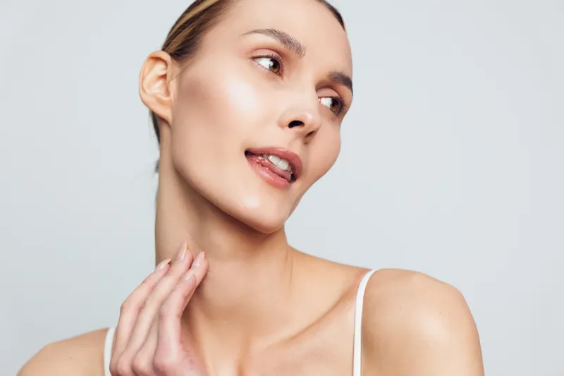 Young woman with clear skin touches her neck gently looks away with natural expression on light background wearing white thin strap top minimal makeup healthy beauty concept.