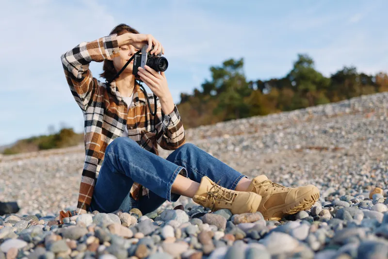 Photographer with camera in hand, kneeling on a pebble beach, focusing through the lens in casual attire, capturing an outdoor moment amid rugged shore and open sky.