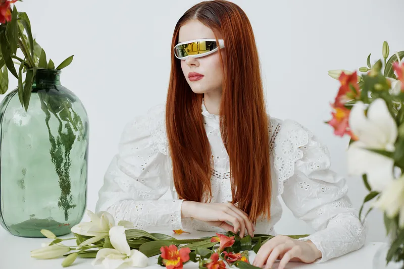 Stylish young woman with long red hair wearing futuristic sunglasses, sitting among fresh flowers and greenery, showcasing a modern aesthetic and a serene expression