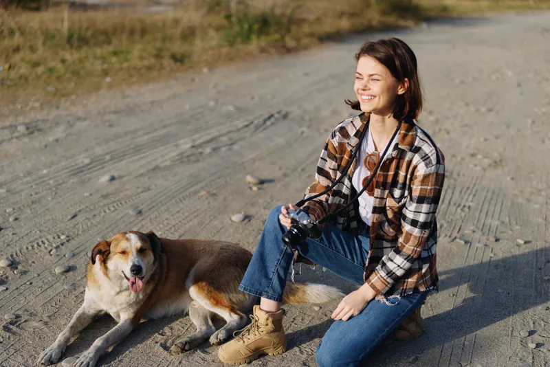 Woman kneeling beside a dog on a dusty road, outdoor casual scene with a plaid shirt, jeans and boots, joyful expression, field setting and warm sunlight.