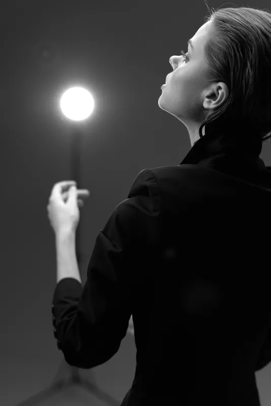 Black and white portrait of a woman holding a glowing orb with a contemplative expression in a minimalistic setting.