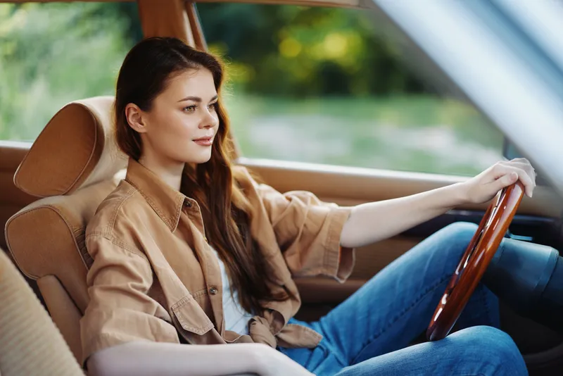 Young woman in a stylish beige outfit, sitting in a vintage car, smiling and enjoying the drive in a scenic outdoor background.