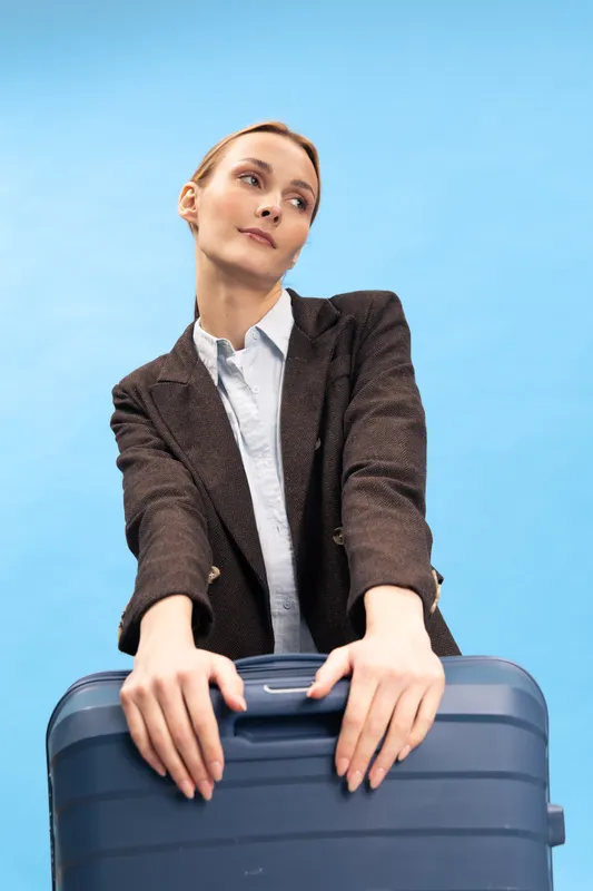 Young woman in brown blazer stands holding a blue suitcase against a clear blue sky. Concept of travel, business trip, and planning. Casual confident female traveler in daylight outdoors.