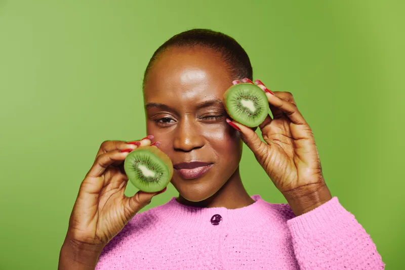 Close-up of a smiling black woman with dark skin holding two kiwi halves near her face on a green background. Concept of healthy lifestyle, natural food, and beauty care.