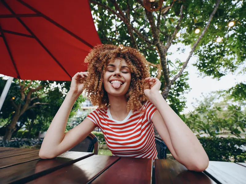 Young woman with curly hair sits at an outdoor wooden table under a red umbrella, wearing a striped shirt and sticking her tongue playfully toward the camera.