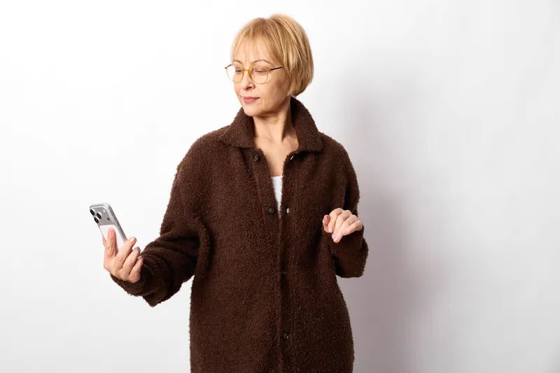 Elderly woman with glasses, holding a smartphone indoors, wearing a dark coat, focused on the screen, calm communication gesture, clean white background, soft diffused light