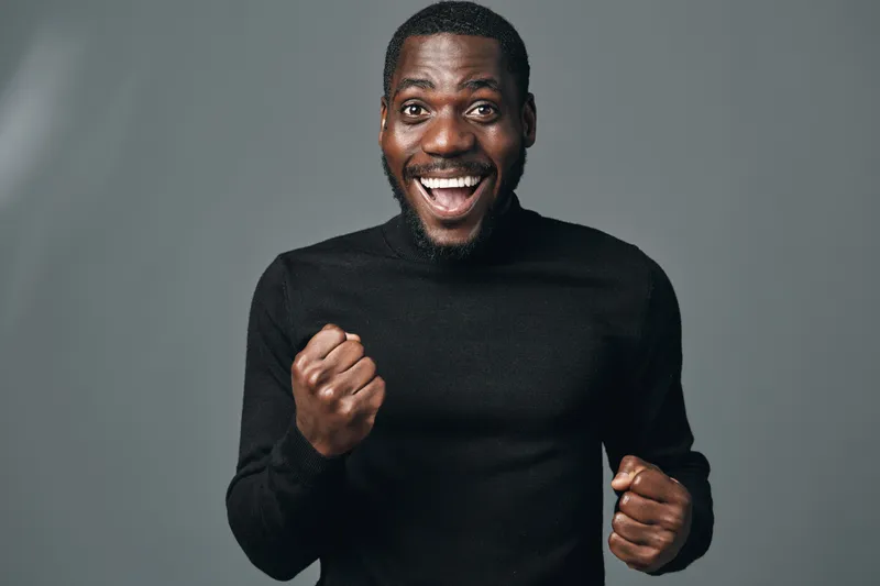 African man with dark skin wearing black sweater showing excitement and joy with clenched fists and wide smile in creative conceptual visual style portrait on gray background.