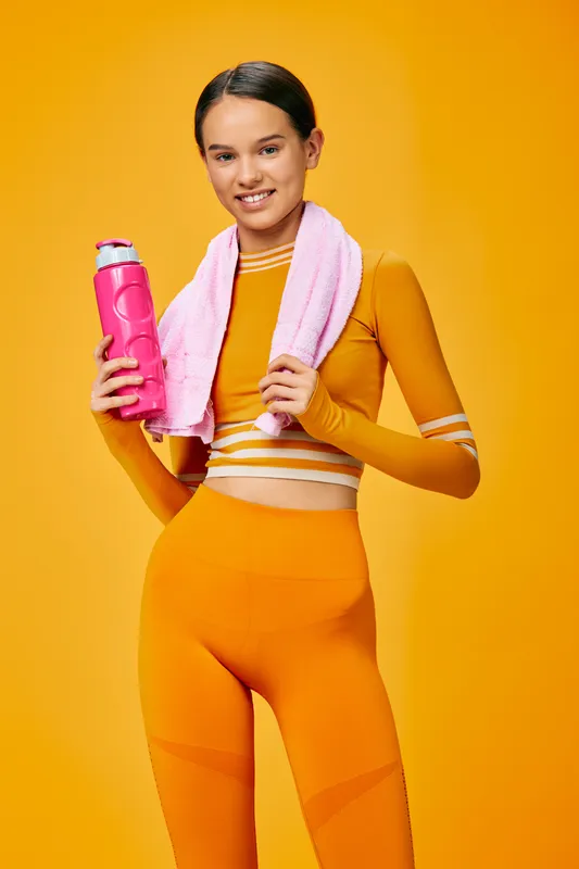 Female athlete in bright orange activewear with a pink water bottle and towel, posing confidently after a workout against a vivid orange backdrop, showcasing health, strength and motivation in a