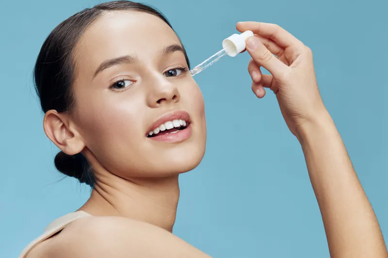 Young woman applying serum on her face with a dropper, showcasing radiant skin and a happy expression, against a soft blue background