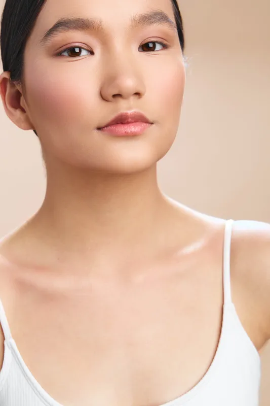 Asian woman gazes softly into the camera with a calm expression, wearing a white tank top. The close up portrait highlights smooth skin and subtle makeup, conveying serenity and confidence in a
