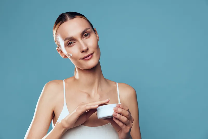 Young woman with brunette hair applies moisturizing cream on her hand while posing in front of a blue background with a gentle smile.