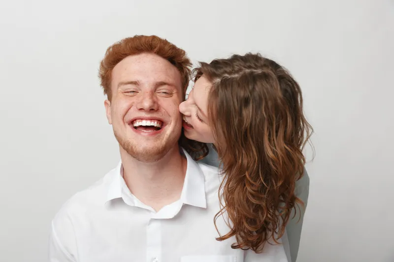 Happy young couple in casual clothes share a joyful moment, woman kissing man on cheek. White background, natural light, concept of love, affection, friendship, and happiness.