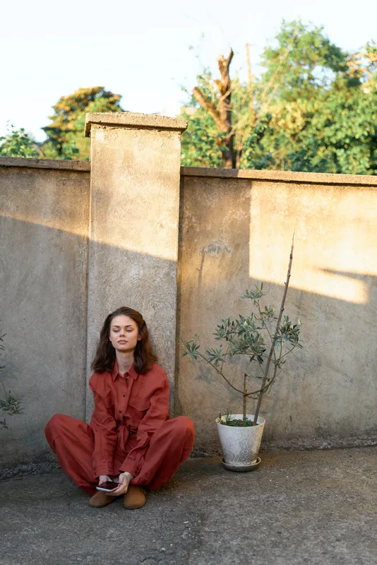 A woman in a red jumpsuit sits on a concrete rooftop, leaning against a wall with eyes closed, conveying calm solitude and urban lifestyle, sunlit backdrop