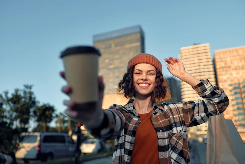 Woman holding coffee cup to camera, smiling in urban street wearing beanie and carrying shopping bag, candid moment with authenticity, golden hour glow and mindful living vibe.