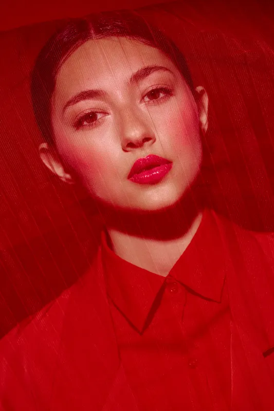 Close-up of a young woman with flawless skin wearing vibrant red lipstick and a red shirt standing behind red fabric. Creative fashion and beauty concept with strong red tone and mood.