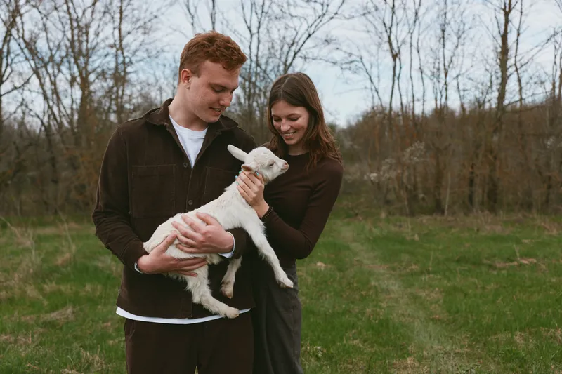A young couple enjoys a tender moment outdoors as the man holds a white baby goat while the woman gently touches it in a natural spring setting with bare trees.