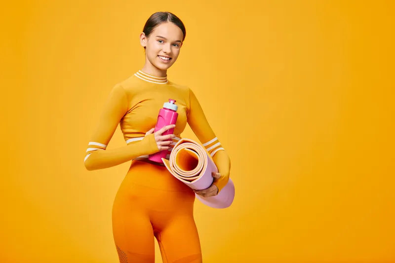 Vibrant image of a woman in bright orange athletic wear holding a yoga mat and water bottle against a bold orange backdrop, ready for a dynamic fitness session.