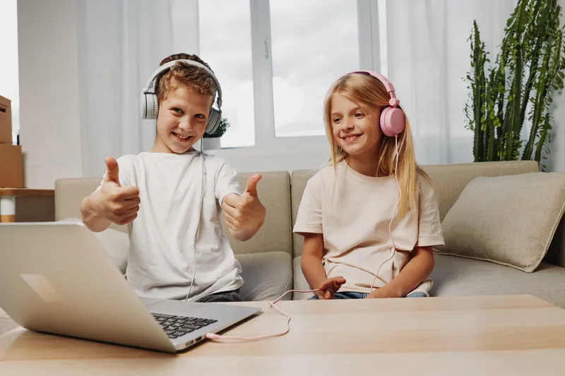 Happy children wearing headphones enjoy music and use laptop on sofa in bright room. Concept of technology, entertainment, childhood, and digital learning at home with casual clothes and relaxed mood.