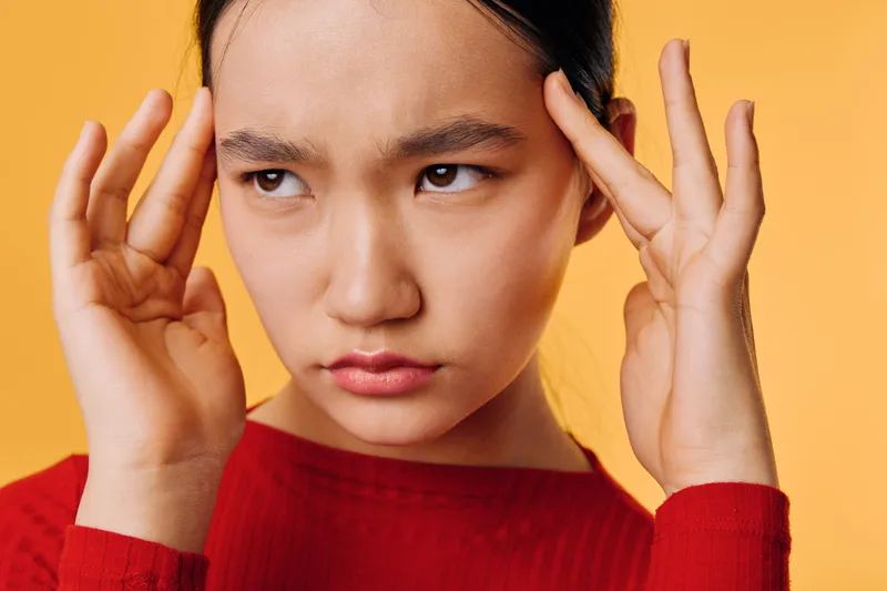 Young woman with serious expression touching temples and frowning with concentration on orange background. Closeup portrait, Asian female with dark hair wearing red sweater.