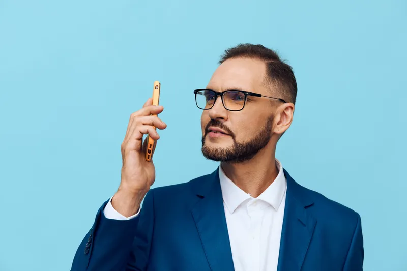 A focused, confident business professional speaks on a phone in a blue suit against a solid blue background, conveying attentive mood, calm determination, and strong presence in a studio setting.