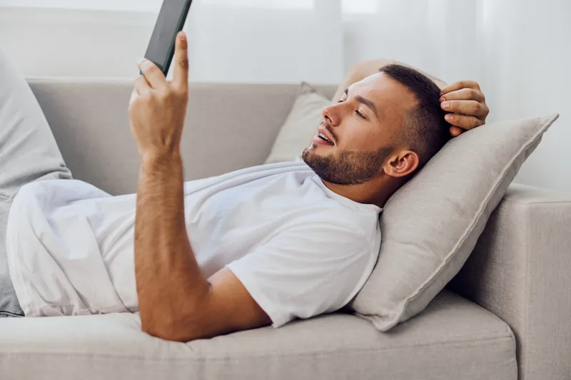 Engaged man relaxing on a sofa, using a tablet with a joyful expression, cozy interior setting. Perfect for lifestyle themes and modern technology concepts.