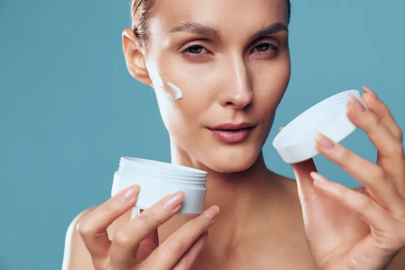 Young woman applies moisturizing cream on face holds white jar with cosmetic product looks at camera with soft natural makeup isolated on blue background skincare concept.