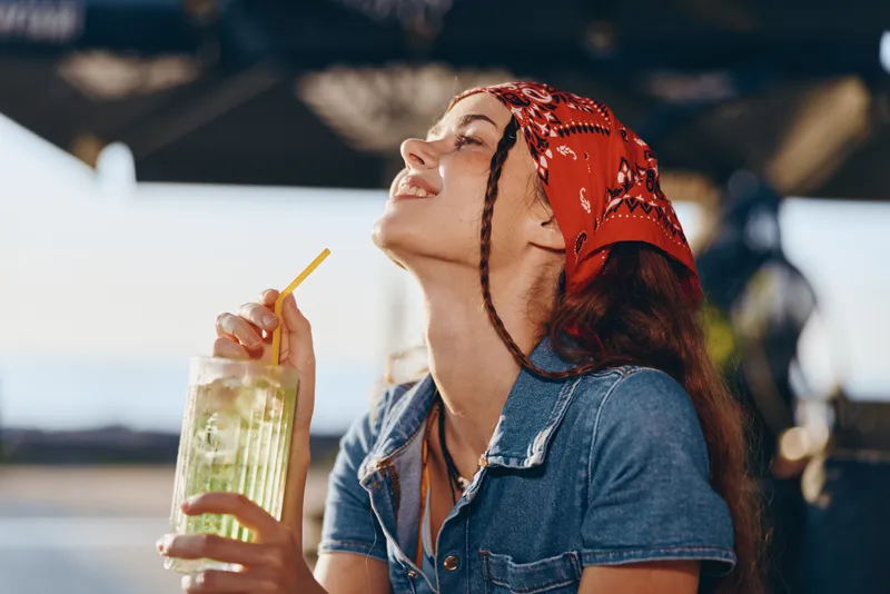 Lifestyle woman in modern boho-western denim street style enjoys refreshing drink with straw, smiling and relaxed outdoors under warm film color tones.