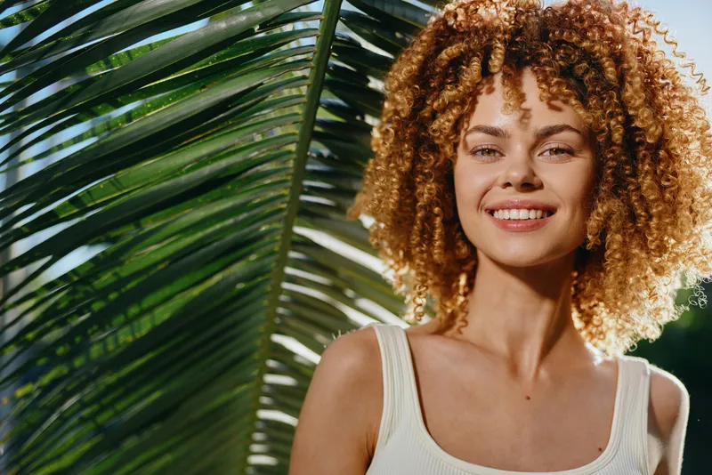 Smiling woman with curly hair stands near green palm leaf outdoors, wearing a white tank top, basking in natural sunlight with a joyful expression on her face.
