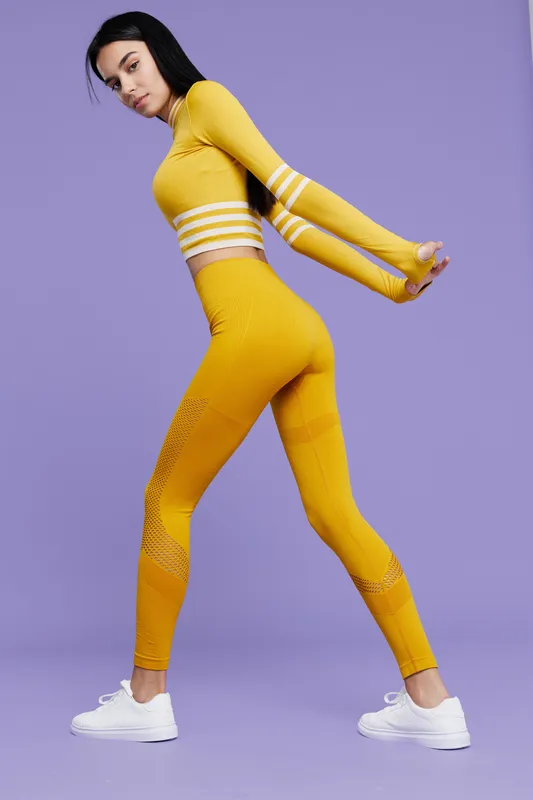 Woman sport fitness stretching in bright yellow activewear and white sneakers, preparing for workout on purple background, showcasing flexibility and athleticism.