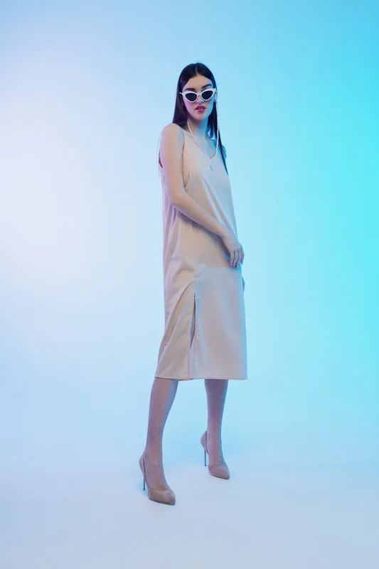 Young woman in a stylish beige dress and sunglasses poses confidently on a gradient blue background. Concept of woman beauty fashion, elegance, and modern style.