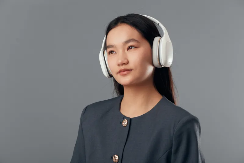Young woman wearing wireless headphones looking thoughtful in studio. Female person with long dark hair enjoying music or podcast with gray background and casual clothing.