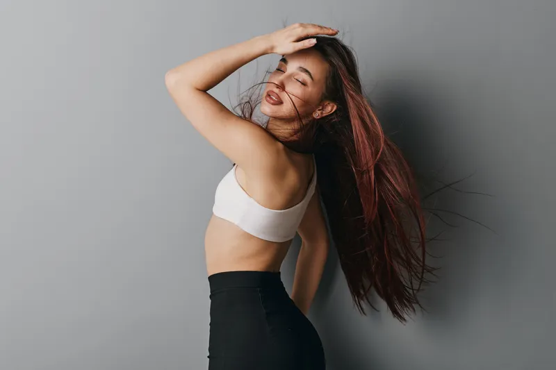 Young woman with long hair wearing sports bra and leggings posing with eyes closed and hand on head against gray background for fitness and beauty concept.