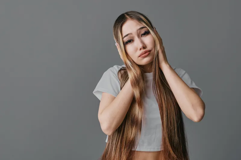 Young woman with long braided hair wearing white crop top touching her head and showing worried facial expression on gray background in studio.