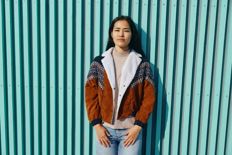 Casual street style featuring a colorblock knitted cardigan over a light turtleneck, standing against a turquoise striped wall for a fashion photo session with urban charm and confident presence