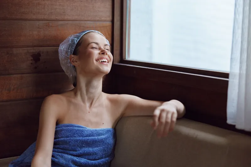 smiling woman wrapped in blue towel relaxing indoors near window with natural light and wooden wall background, feeling peaceful and refreshed