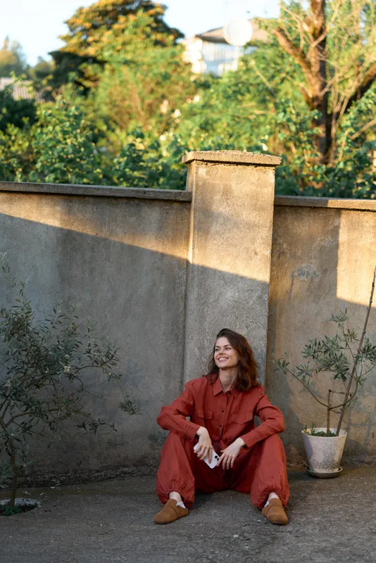 Woman in a red jumpsuit sits on the ground beside a wall in a garden, smiling softly, capturing a casual lifestyle moment outdoors with relaxed pose, warm sunlight, candid vibe, and natural textures