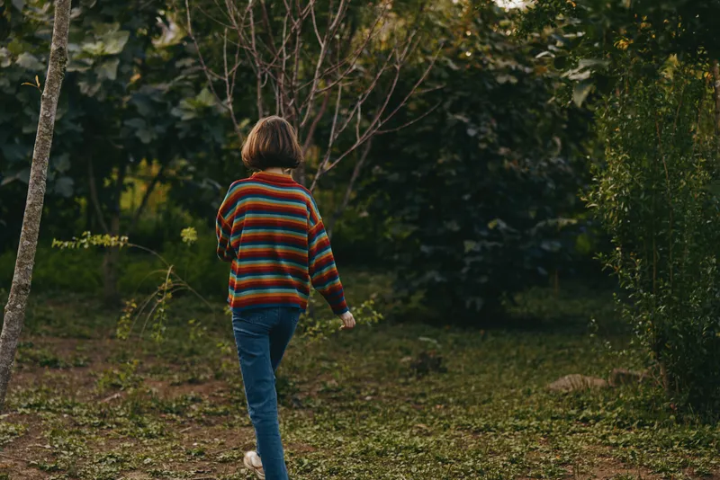 Child girl walking in a garden nature scene wearing a striped sweater, back view outdoors in autumn casual outfit exploring greenery and trees along a grassy path.