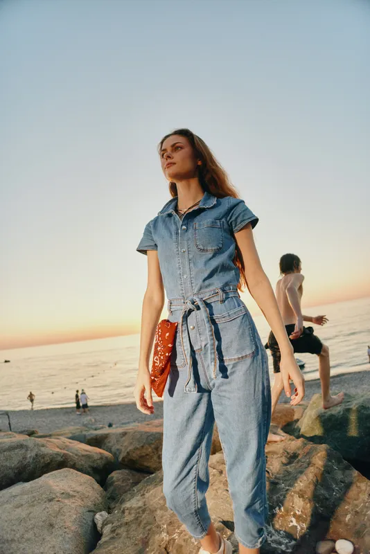 young woman in denim jumpsuit walking on rocky beach at sunset with calm sea and warm light, casual style and thoughtful expression
