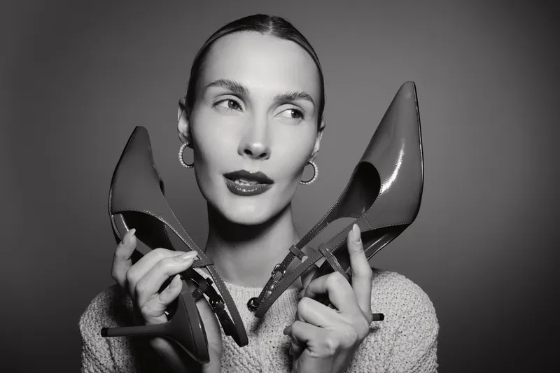 Young white woman with light skin holds high heel shoes near her ears, wears hoop earrings and knitted sweater. Studio portrait in black and white with thoughtful mood.