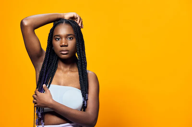 A young Black woman with braided hair poses confidently against a vibrant yellow backdrop, showcasing a fashionable crop top and expressing strong emotions of empowerment and beauty