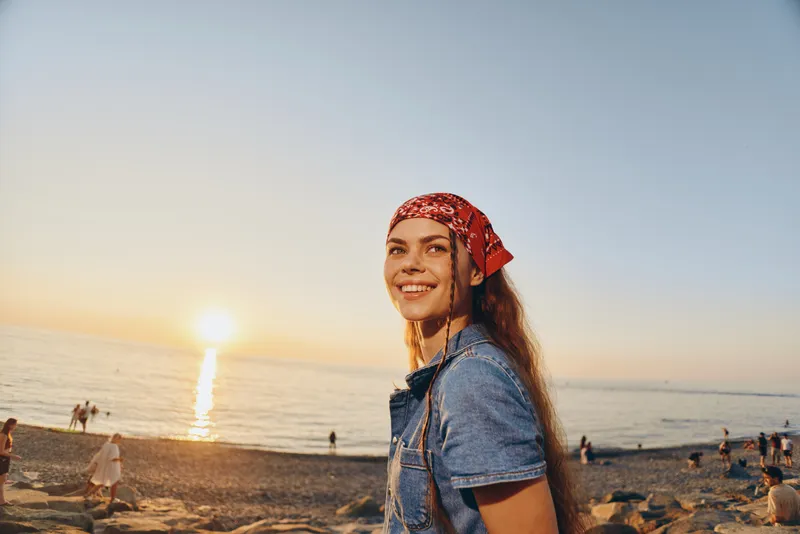 lifestyle woman in modern boho-western denim street style smiling near the beach at sunset with warm film color tones