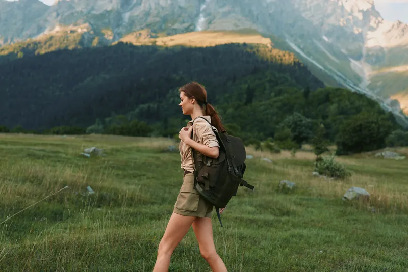 Young woman hiking outdoors with backpack walking through green meadow with mountains in background enjoying nature and active lifestyle on sunny day