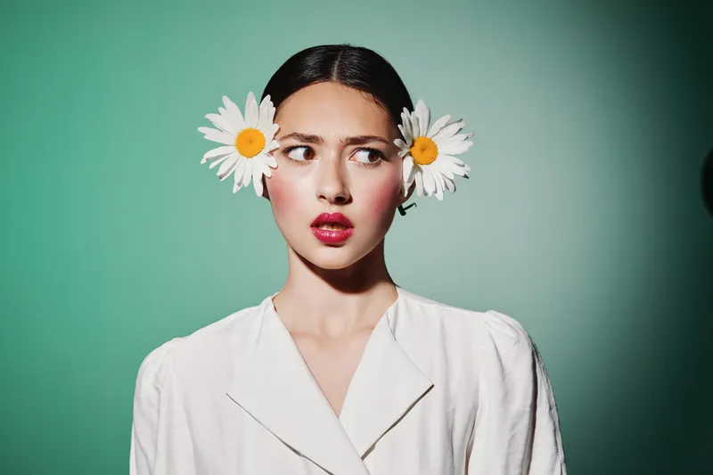 Young woman with white flowers in her hair looks sideways with curiosity. She wears a white blouse against a turquoise background. Concept of creative beauty and fashion styling.