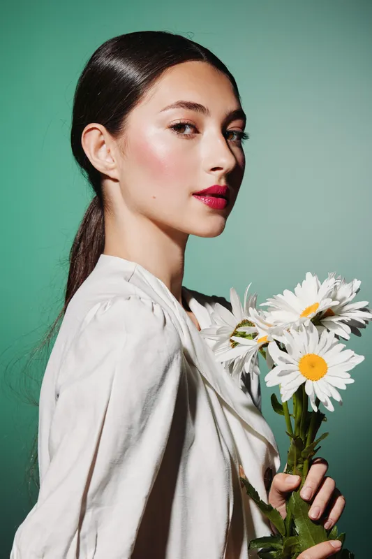 Fashionable woman with sleek hair and radiant makeup poses with white daisies against green backdrop. Creative beauty concept highlighting skincare, floral elements, and elegant style.